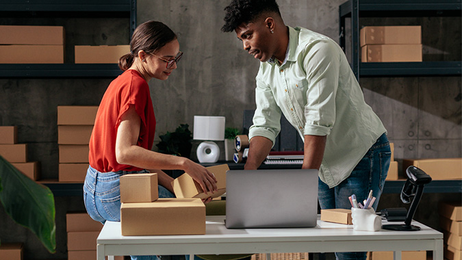 Two people working together at a desk in a workspace or warehouse, with shelves filled with boxes in the background. One person is holding a cardboard box while the other is using a laptop.