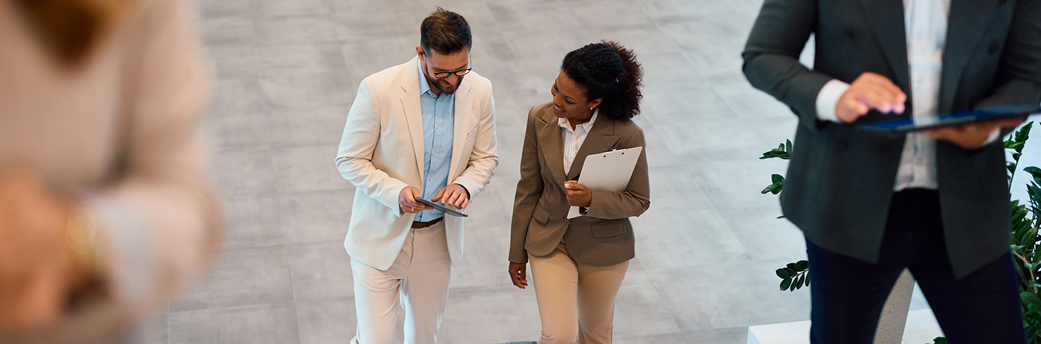 A man and woman look at a tablet device as they walk up the stairs of an office building. 