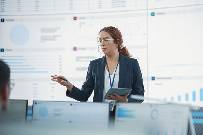 Business professional presenting data on large screens, holding a tablet and a pen, with audience monitors visible in the foreground.