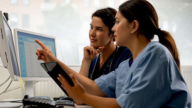 Two nurses sit at a desk looking at computer monitors. One nurse holds a tablet.