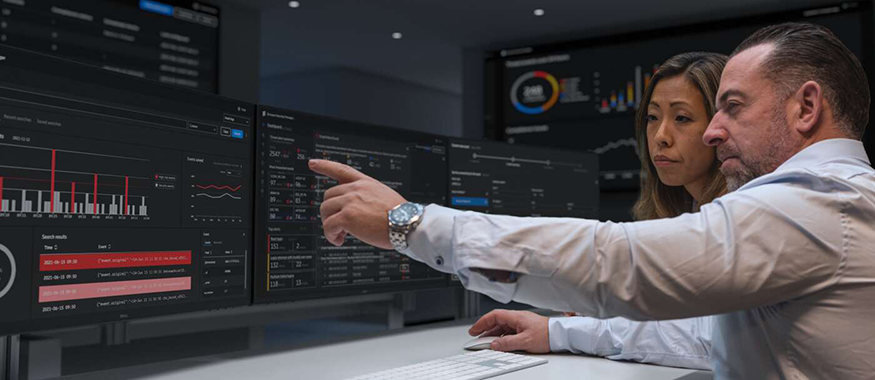 Two people review analytics dashboards on multiple widescreen monitors.