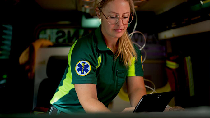 A paramedic in a green uniform works on a tablet while inside an ambulance.