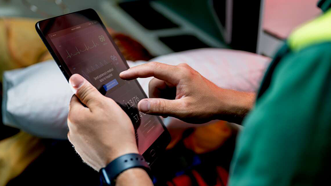 Hands operating a tablet that displays ECG traces and vital signs, monitoring a patient inside an ambulance.