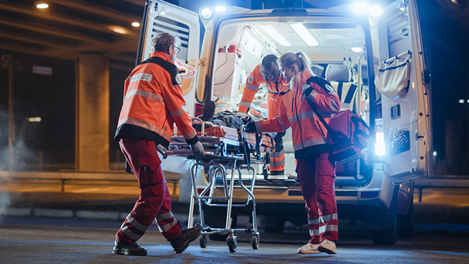 Three emergency medical technicians, wearing high-visibility orange jackets and red pants, load a patient on a stretcher into the brightly lit back of an ambulance at night under an overpass.