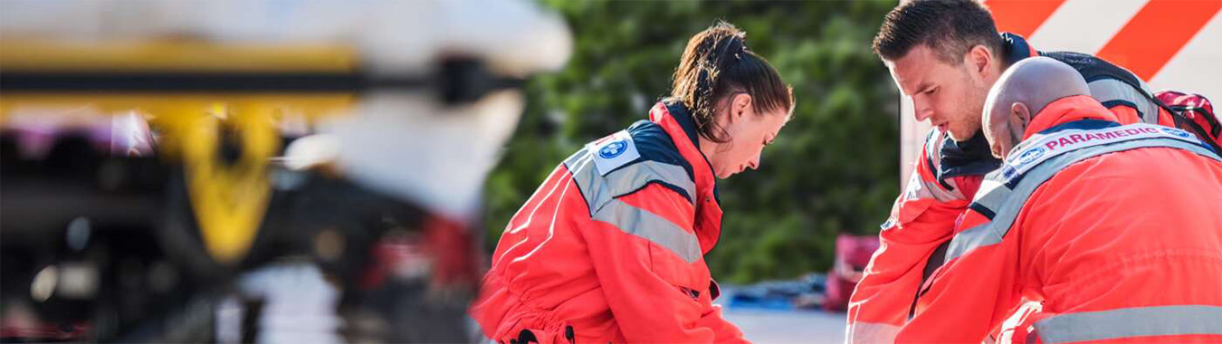 Three paramedics in bright orange uniforms are attending to a patient on the ground, with medical equipment nearby and an emergency vehicle in the background.