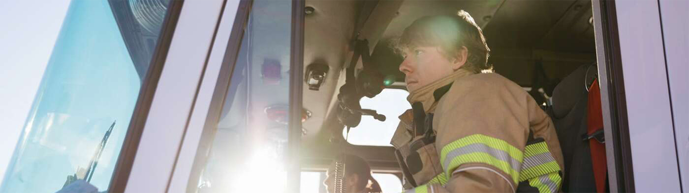 A firefighter in protective gear is seated inside a fire truck, speaking into a radio while sunlight streams through the window.