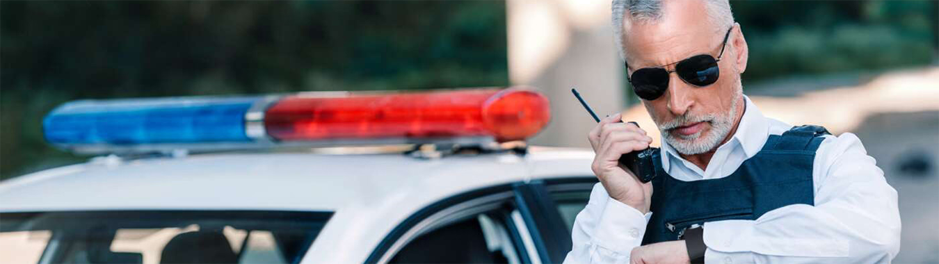 Police officer in a bulletproof vest speaks into a radio while standing beside a patrol car with red and blue lights.