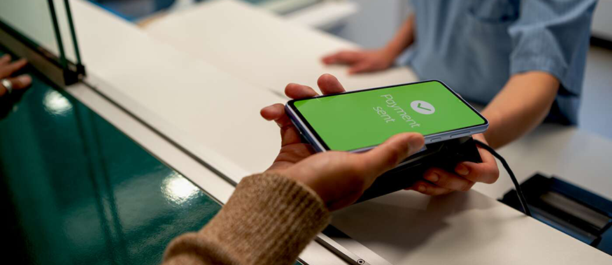 Close-up of a contactless mobile payment at a reception desk—smartphone screen shows 'Payment sent' with a checkmark as a staff member in blue scrubs stands behind the counter.